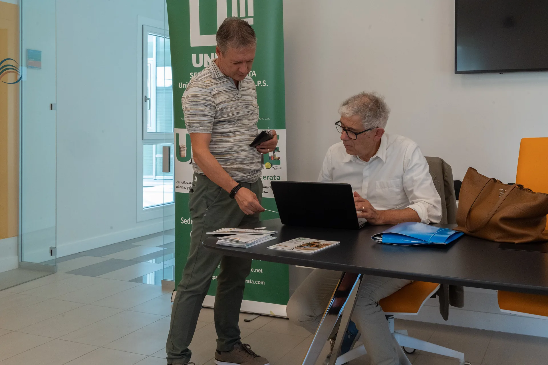 Due uomini al tavolo con il portatile e i materiali informativi di Unitre, davanti al roll-up